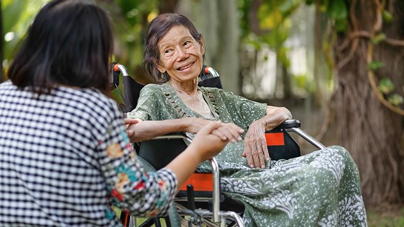Elderly woman sitting in wheelchair in green dress smiling and holding hands with younger woman with her back to camera