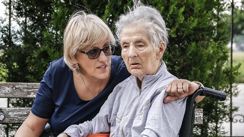 Woman in sunglasses with arm around wheelchair speaking to elderly woman in wheelchair in outdoor setting