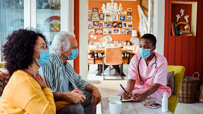 Caregiver in home setting wearing mask talking to couple wearing masks at coffee table